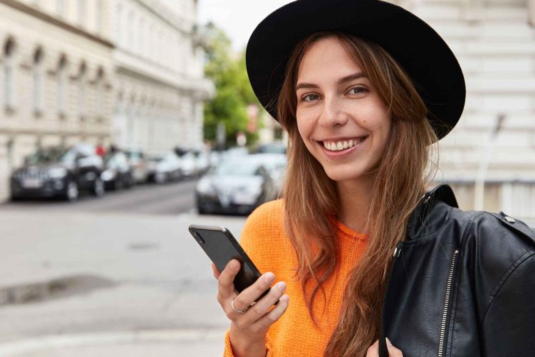 Eine lächelnde junge Frau mit Hut, die ihr Handy in der Hand hält.