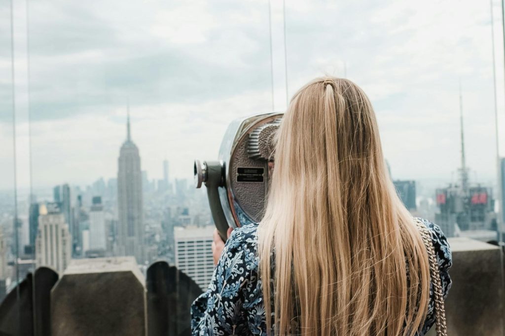 Eine junge Frau mit langen blonden Haaren vor einem Panorama-Fernglas am Top of the Rock in New York.