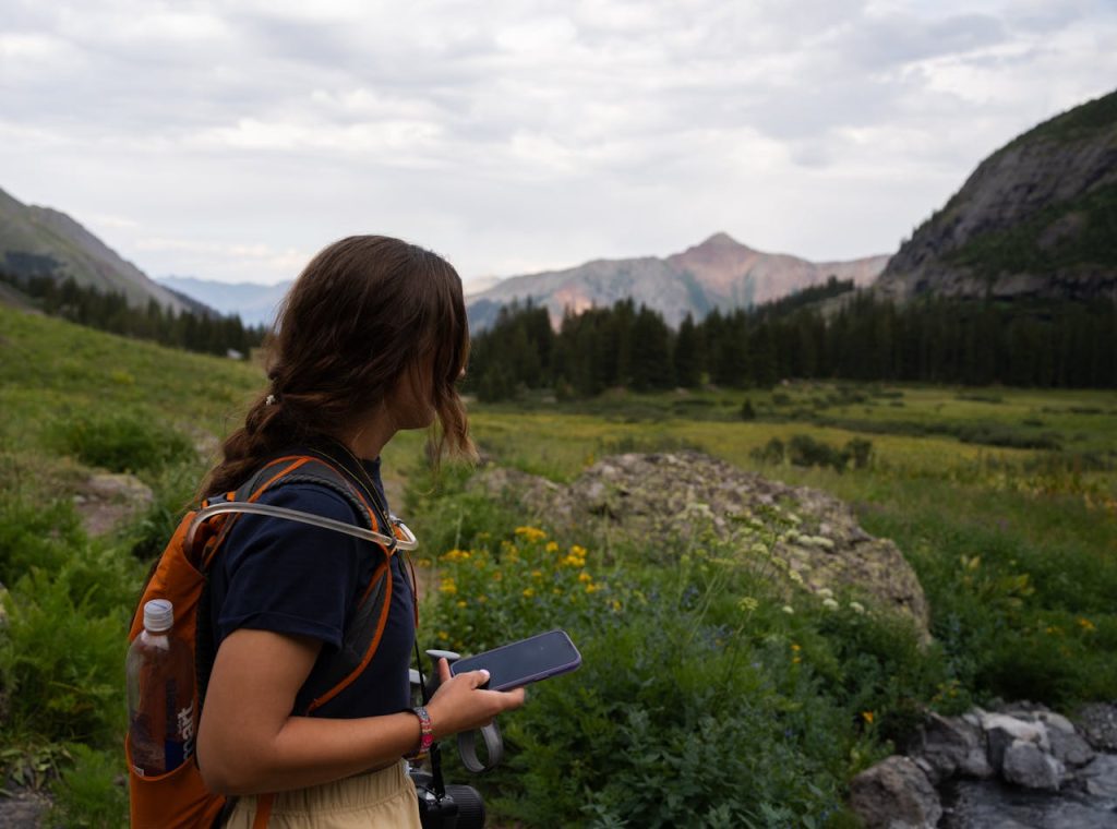 Eine Frau mit einem Rucksack, die in einer nordamerikanischen Mittelgebirgslandschaft nach einem Handy Ausschau hält.