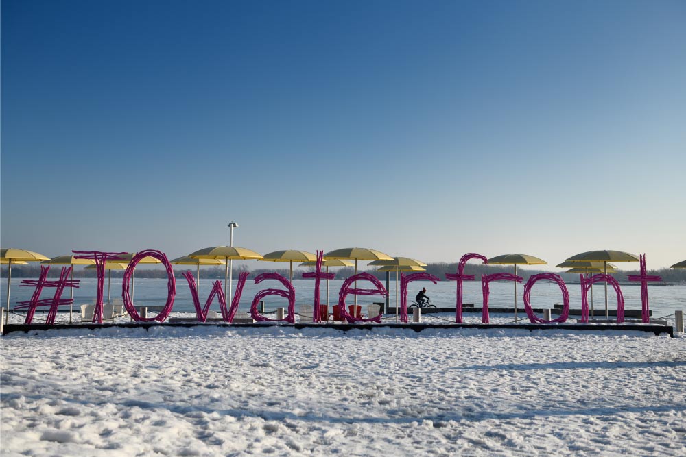 雪に覆われたトロントのウォーターフロントの「#TOwaterfront」の看板。背景には黄色のパラソルと湖が広がっている。