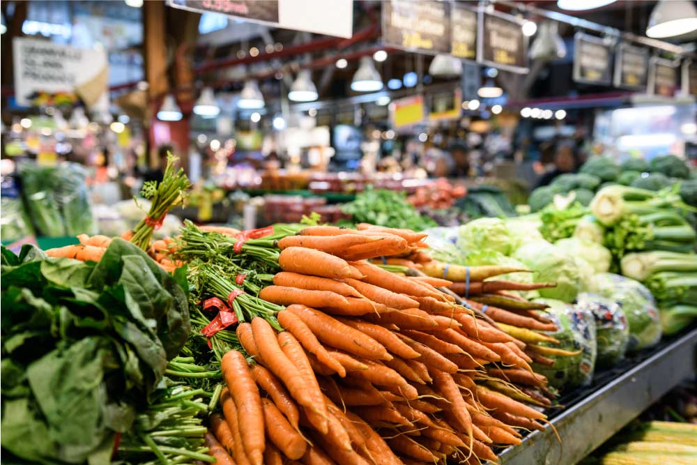 グランビルアイランド市場で売られている新鮮な野菜のクローズアップ。市場の賑わいを背景に、カラフルなにんじんやその他の野菜が展示されている。