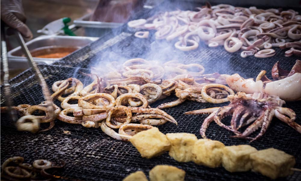 屋台で焼かれているイカの輪切りとゲソ、スモークが立ち上る美味しそうなシーン。