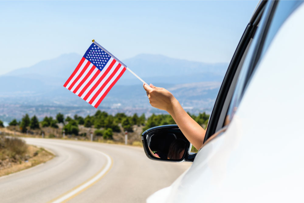 車の窓からアメリカ国旗を持っている手の様子、背景には山や自然が広がる道路。