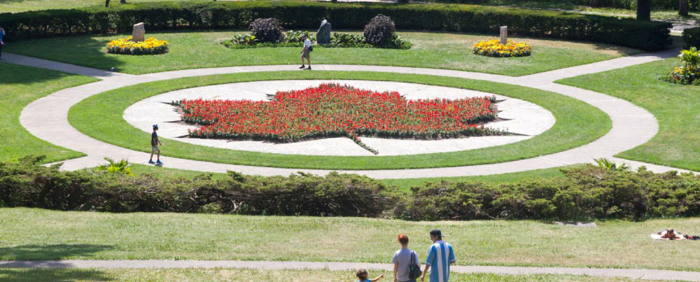 カナダの公園でメープルリーフの形に整えられた花壇。赤い花がメープルリーフの形を描き、訪れる人々が散策している