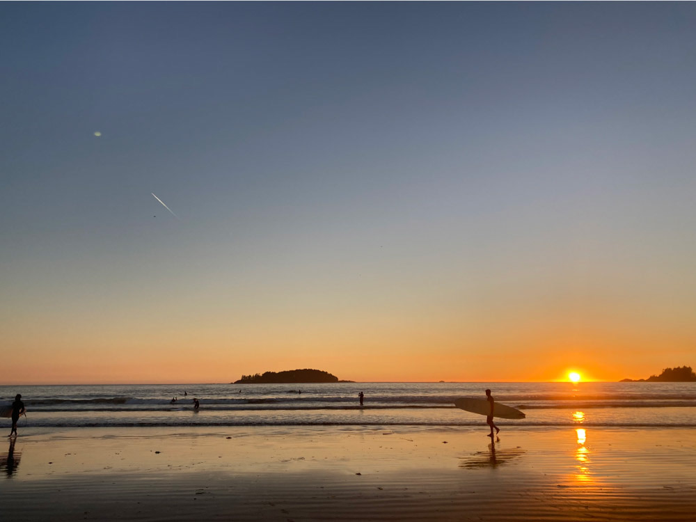 トフィーノのビーチで、夕日を背景にサーフボードを持つサーファーたちが波打ち際に立っているシーン。