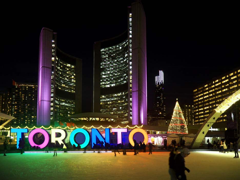 夜のネイサンフィリップススクエアで、クリスマスツリーとイルミネーションに彩られたTORONTOサイン、スケートリンクで楽しむ人々。