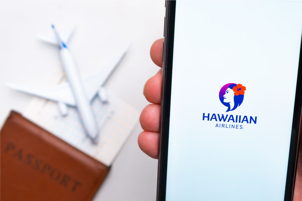 手に持ったスマートフォンに表示されたHawaiian Airlinesのロゴ。背景にパスポートと飛行機の模型が置かれている