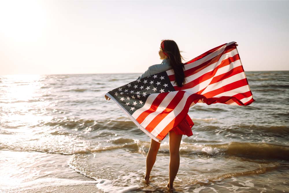 メリカ国旗を持ち、ビーチで夕日を眺める女性