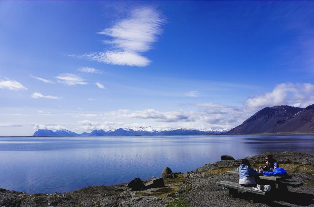 View of a blue sky and blue lake in Iceland, with two people sitting at a table in the foreground and snow-capped mountains in the background. 