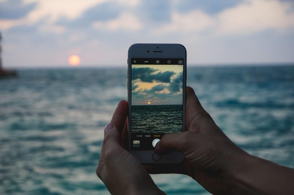 Close up of hands holding a phone taking a picture of the sun setting over the ocean.